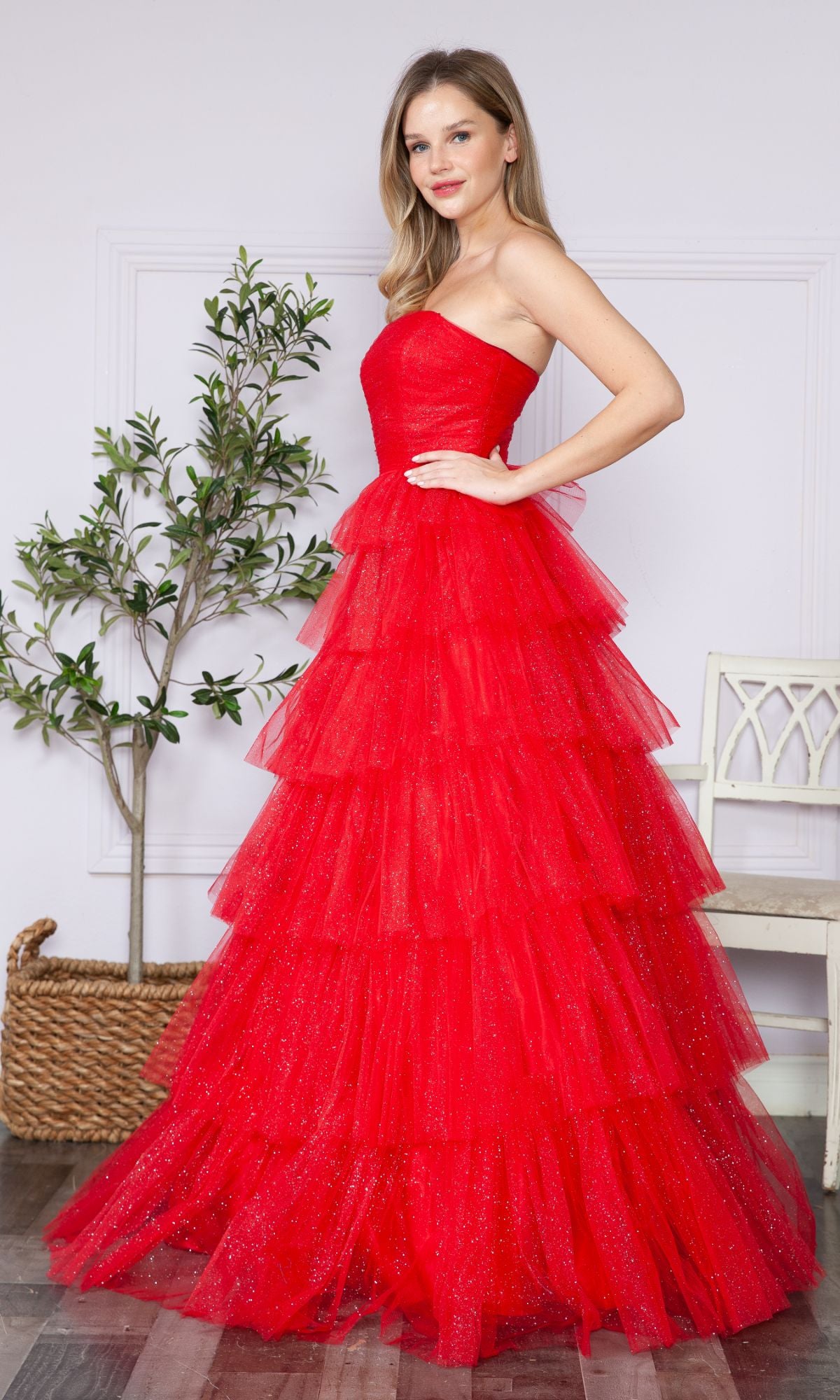 A woman stands indoors beside a potted plant and white chair, wearing the Strapless Long Tiered Glitter Prom Ball Gown 9386—a floor-length red tulle gown with layered ruffles.